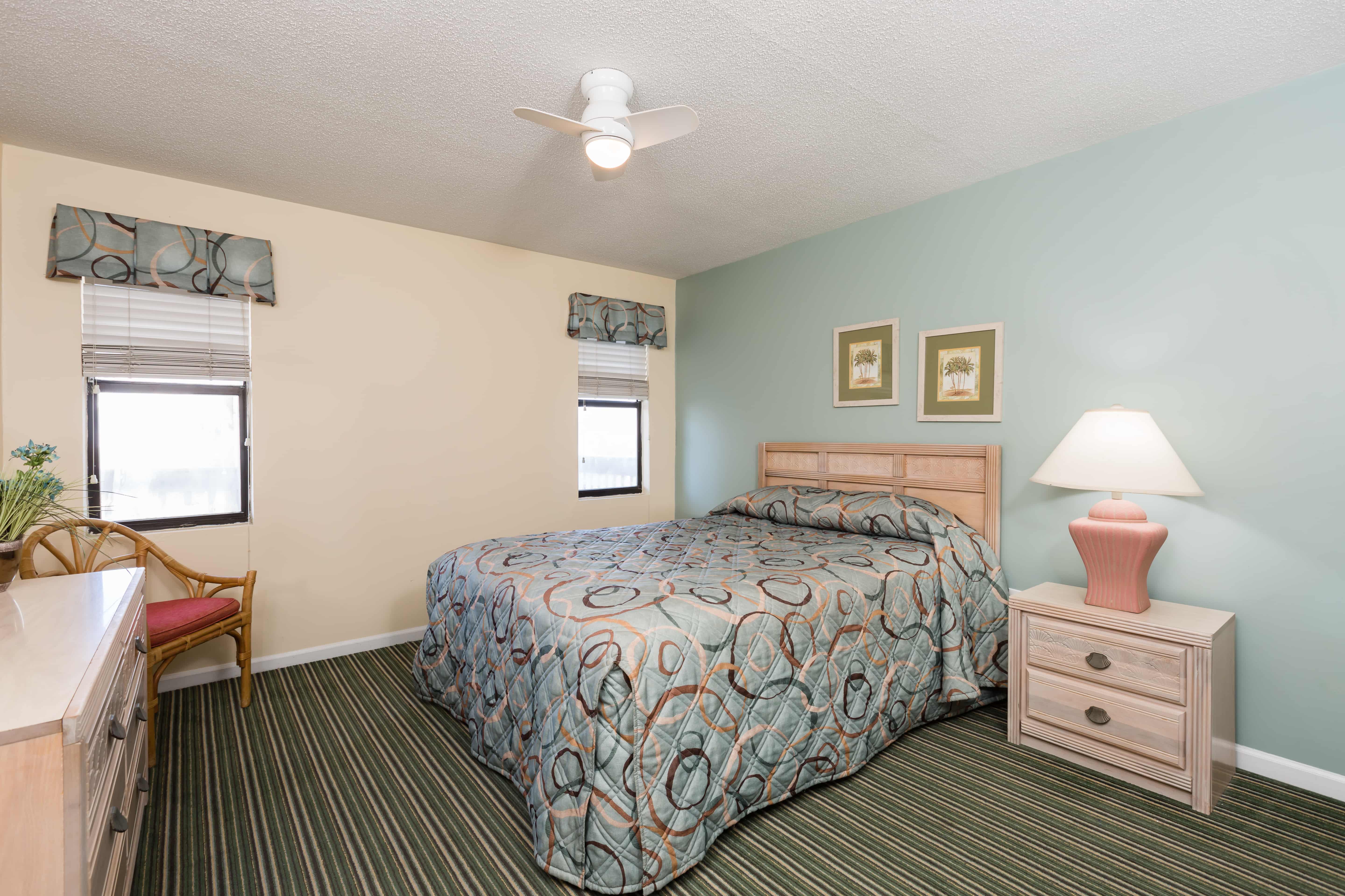 A hotel room with a bed, nightstand, lamp, dresser, chair, ceiling fan, and window