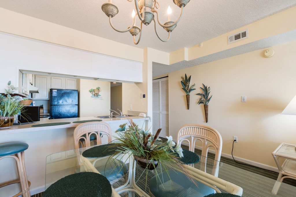 A wide view of a dining area with a glass table and chairs, a kitchenette in the background, and a chandelier hanging from the ceiling.