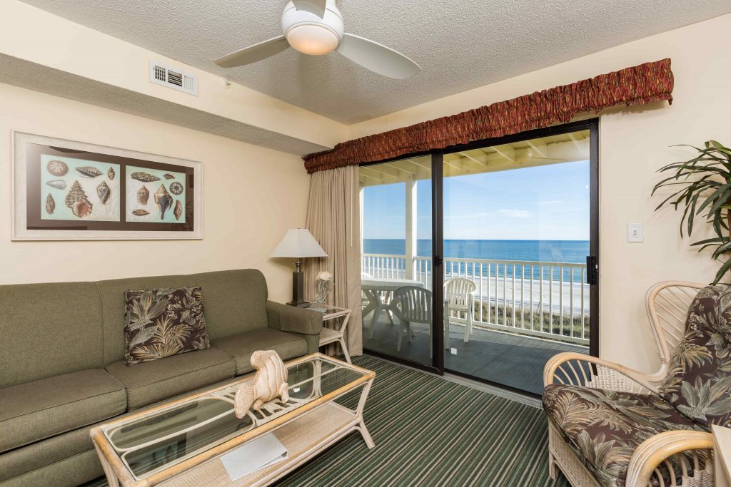 A living room with a sofa, coffee table, chair, and ceiling fan, overlooking the ocean through a sliding glass door.